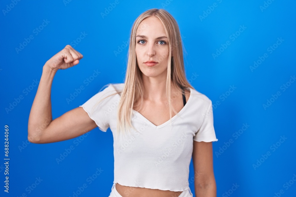 Fototapeta premium Young caucasian woman standing over blue background strong person showing arm muscle, confident and proud of power