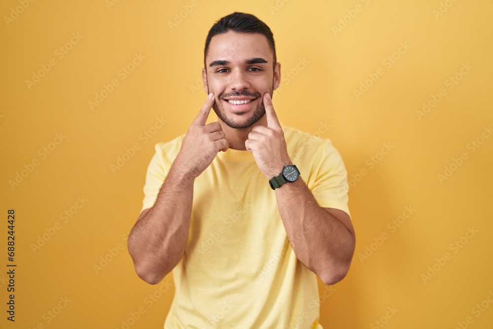 Young hispanic man standing over yellow background smiling with open mouth, fingers pointing and forcing cheerful smile Young hispanic man standing over yellow background smiling with open mouth, fingers pointing and forcing cheerful smile