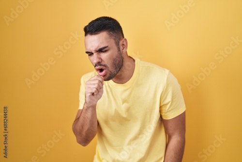 Young hispanic man standing over yellow background feeling unwell and coughing as symptom for cold or bronchitis. health care concept.