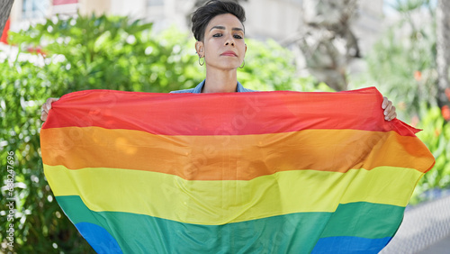 Young beautiful hispanic woman standing with serious expression holding rainbow flag at park
