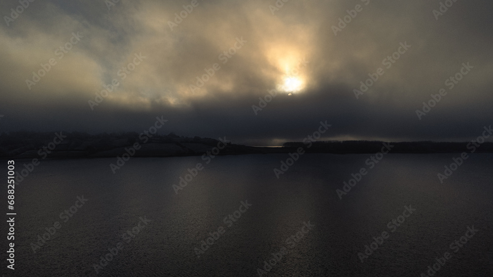 Aerial view of rutland Water reservoir lake as the sun sets in icy conditions with fog over the water and orange trees in late Autumn Fall in Rutland, England UK. 