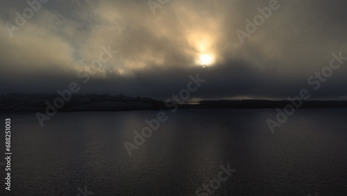 Aerial view of rutland Water reservoir lake as the sun sets in icy conditions with fog over the water and orange trees in late Autumn Fall in Rutland, England UK. 