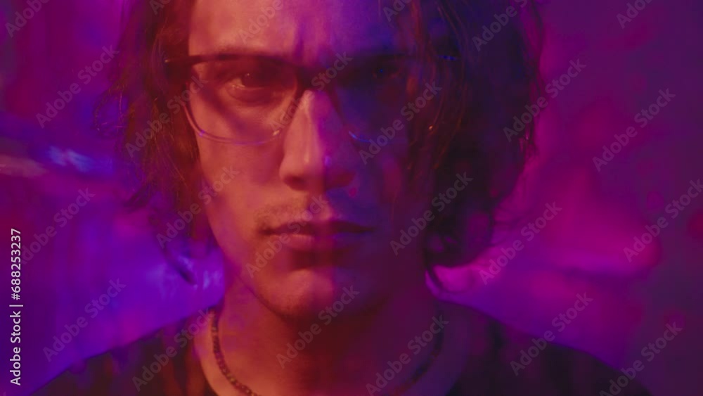 Zoom shot of young man standing in neon light against animated background projection and looking at camera behind glass wall with water droplets running down Zoom shot of young man standing in neon light against animated background projection and looking at camera behind glass wall with water droplets running down