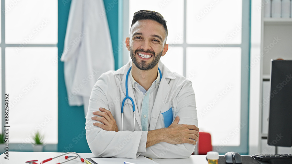 Young hispanic man doctor sitting on table with arms crossed gesture smiling at the clinic