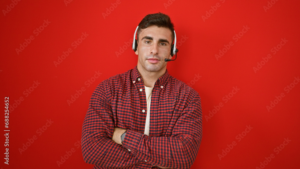 Obraz premium Charming young hispanic man standing firm as call center agent boss, radiating success with relaxed gesture, arms crossed, wearing headset over isolated red wall