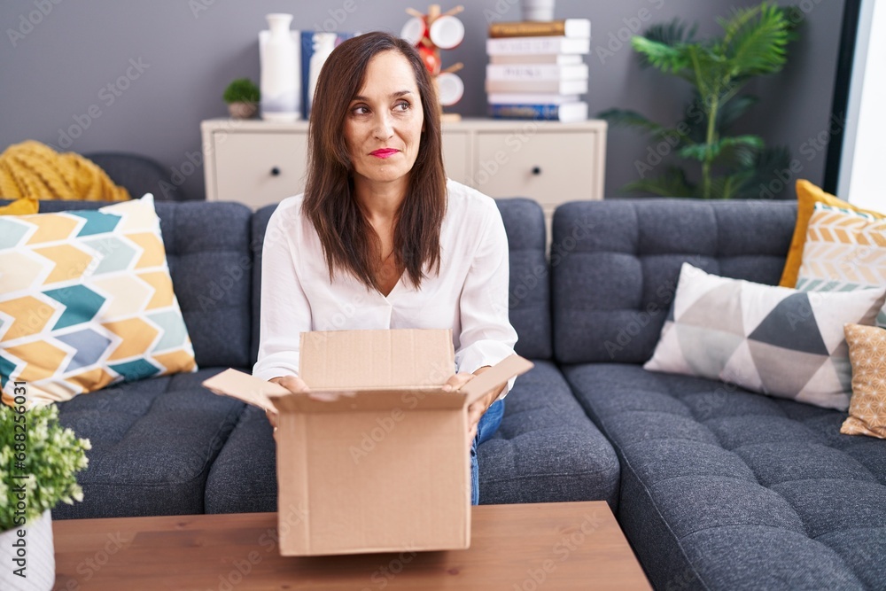 Middle age brunette woman opening cardboard box smiling looking to the side and staring away thinking. Middle age brunette woman opening cardboard box smiling looking to the side and staring away thinking.