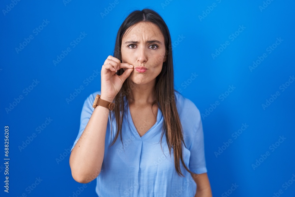 Young brunette woman standing over blue background mouth and lips shut as zip with fingers. secret and silent, taboo talking Young brunette woman standing over blue background mouth and lips shut as zip with fingers. secret and silent, taboo talking
