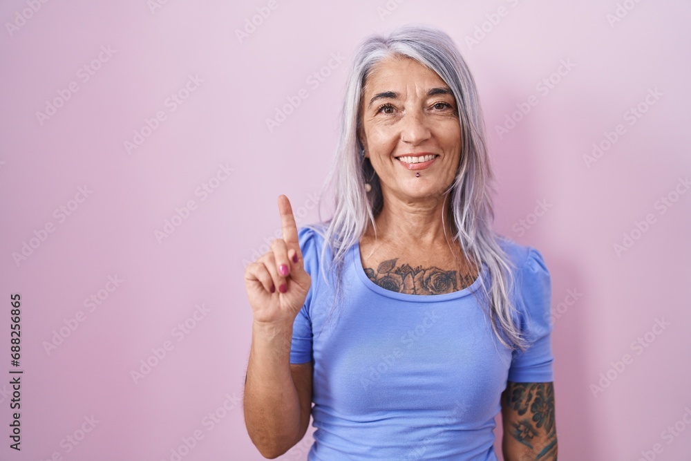 Middle age woman with tattoos standing over pink background showing and pointing up with finger number one while smiling confident and happy.