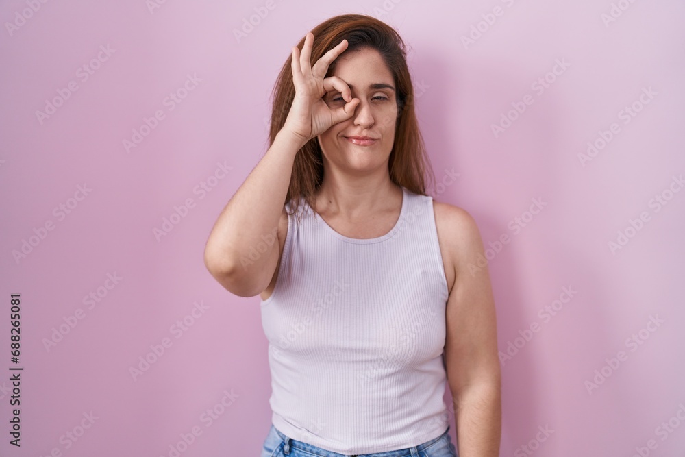 Brunette woman standing over pink background doing ok gesture with hand smiling, eye looking through fingers with happy face. Brunette woman standing over pink background doing ok gesture with hand smiling, eye looking through fingers with happy face.