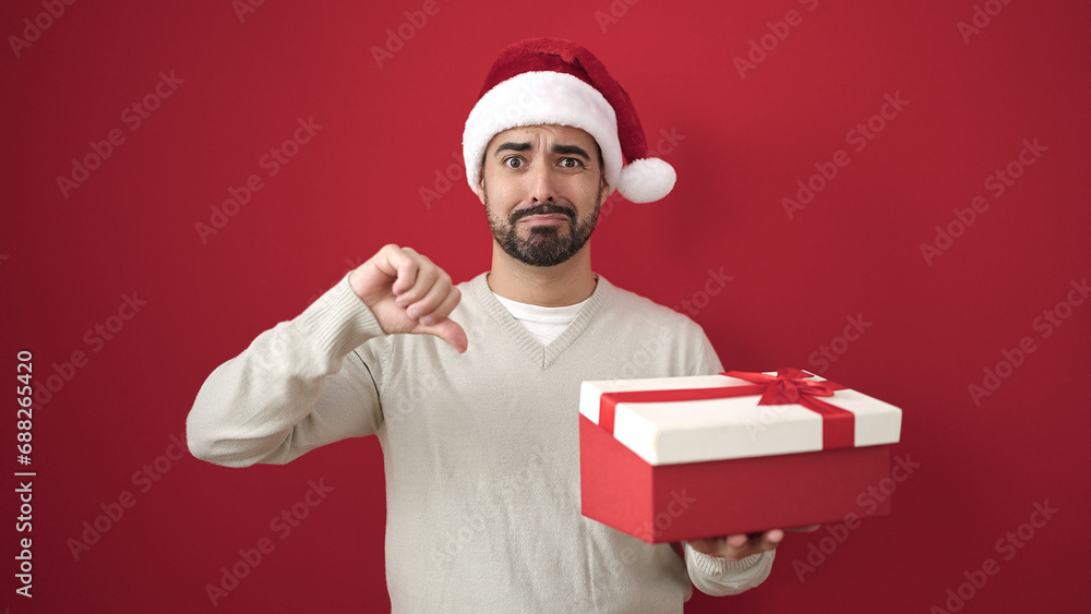 Young hispanic man wearing christmas hat holding gift with thumb down over isolated red background Young hispanic man wearing christmas hat holding gift with thumb down over isolated red background