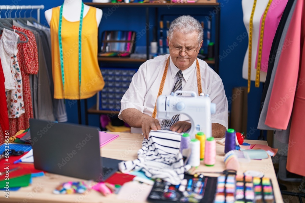 Middle age grey-haired man tailor using sewing machine and laptop at tailor shop Middle age grey-haired man tailor using sewing machine and laptop at tailor shop