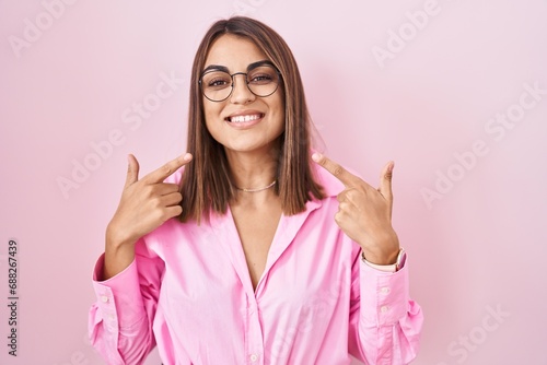 Young hispanic woman wearing glasses standing over pink background smiling cheerful showing and pointing with fingers teeth and mouth. dental health concept.