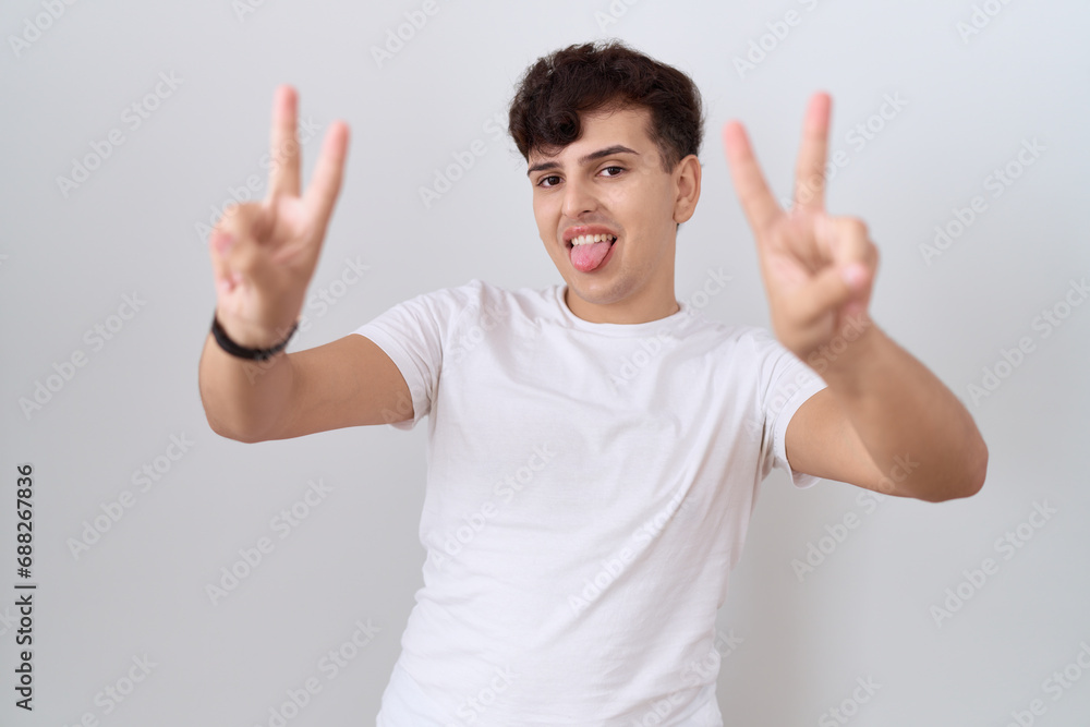 Young non binary man wearing casual white t shirt smiling with tongue out showing fingers of both hands doing victory sign. number two. Young non binary man wearing casual white t shirt smiling with tongue out showing fingers of both hands doing victory sign. number two.