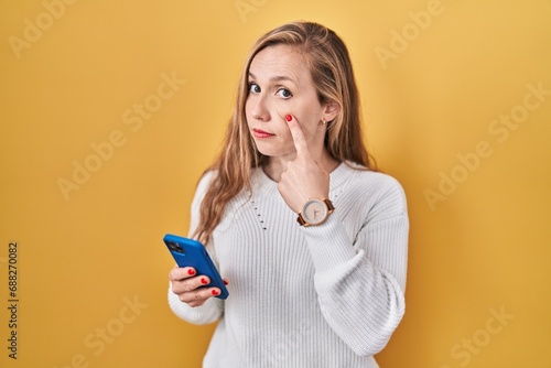 Young blonde woman using smartphone typing message pointing to the eye watching you gesture, suspicious expression photo