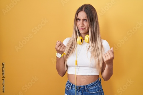 Young blonde woman standing over yellow background wearing headphones doing money gesture with hands  asking for salary payment  millionaire business