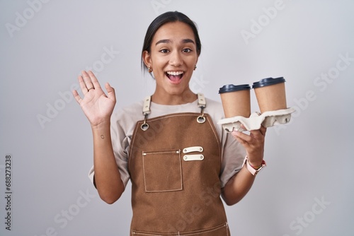 Young hispanic woman wearing professional waitress apron holding coffee waiving saying hello happy and smiling, friendly welcome gesture