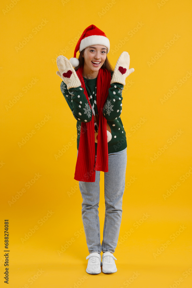 Happy young woman in Christmas sweater, Santa hat and knitted mittens on orange background Happy young woman in Christmas sweater, Santa hat and knitted mittens on orange background