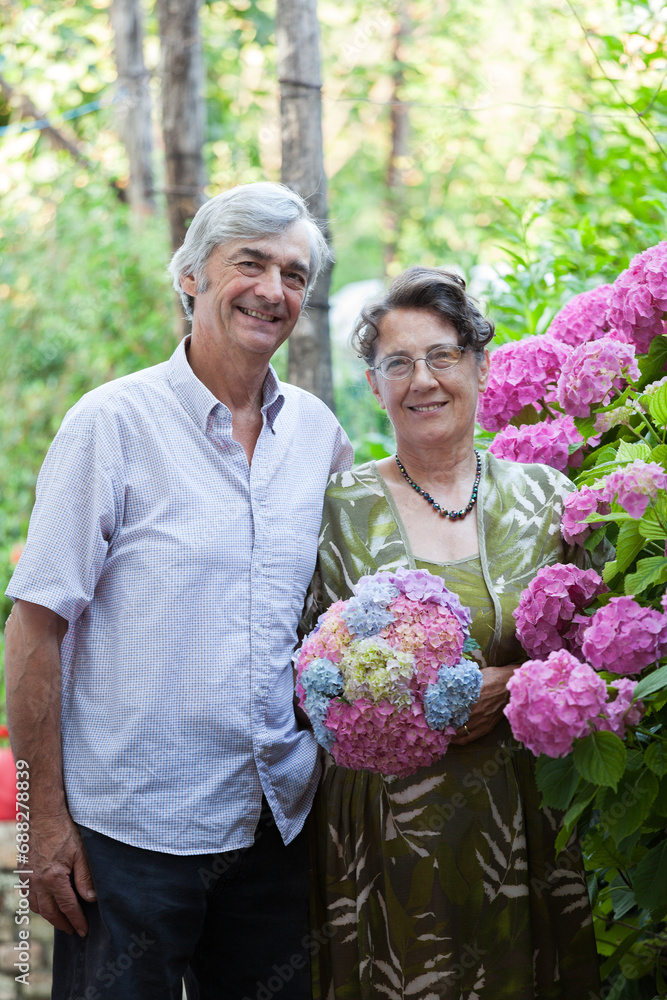 Senior White People Couple, Celebrating 40+ Years of True Love. Genuine Smiles in a Garden Posing for Timeless Togetherness with Colorful Flowers.