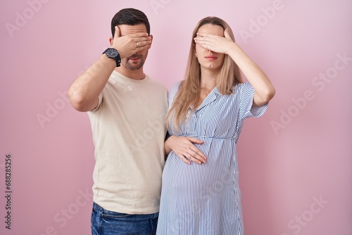 Young couple expecting a baby standing over pink background covering eyes with hand, looking serious and sad. sightless, hiding and rejection concept