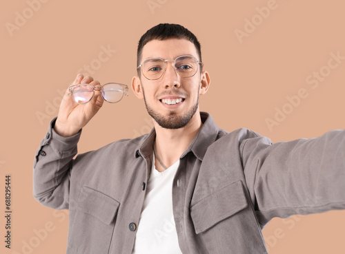 Young man with eyeglasses taking selfie on beige background, closeup