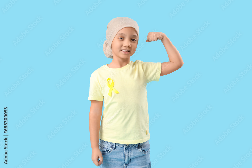 Little Asian girl after chemotherapy with ribbon showing muscles on blue background. Childhood cancer awareness concept Little Asian girl after chemotherapy with ribbon showing muscles on blue background. Childhood cancer awareness concept