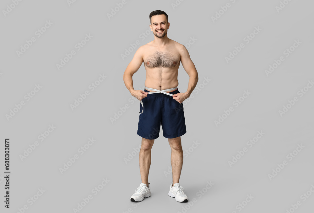 Handsome sporty young man measuring his waist on grey background. Weight loss concept Handsome sporty young man measuring his waist on grey background. Weight loss concept