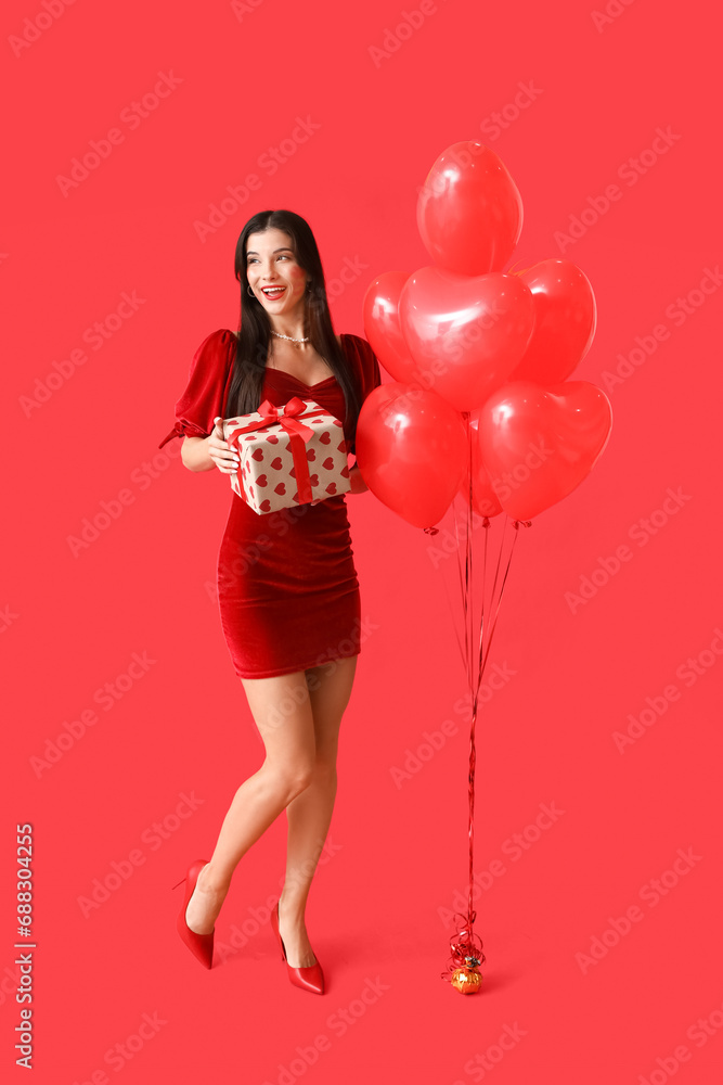 Happy young woman with kiss marks on her face holding gift box and heart shaped air balloons on red background Happy young woman with kiss marks on her face holding gift box and heart shaped air balloons on red background