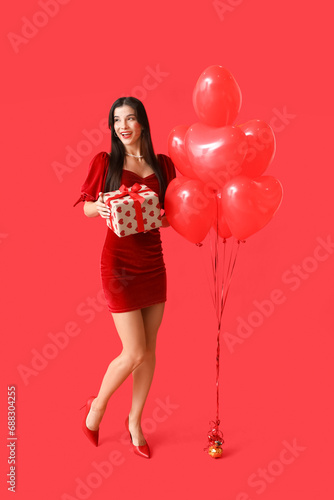 Happy young woman with kiss marks on her face holding gift box and heart shaped air balloons on red background © Pixel-Shot