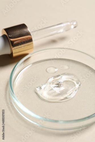Pipette and petri dish with sample of cosmetic serum on beige background, closeup