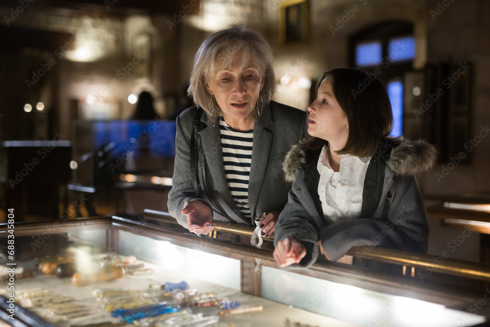 Fototapeta premium Positive senior woman and emotional inquisitive tween girl observing with interest arts and crafts on exhibition in art gallery