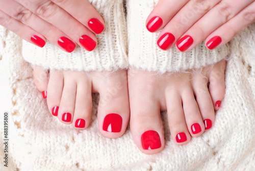 Female feet and hand with red pedicure and manicure on white knitted surface, top view.