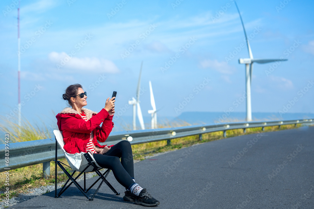 Caucasian woman with red coat and sunglasses sit on chair on roadside near wind turbines or windmill also use mobile phone to take a photo of beautiful view on the mountain.