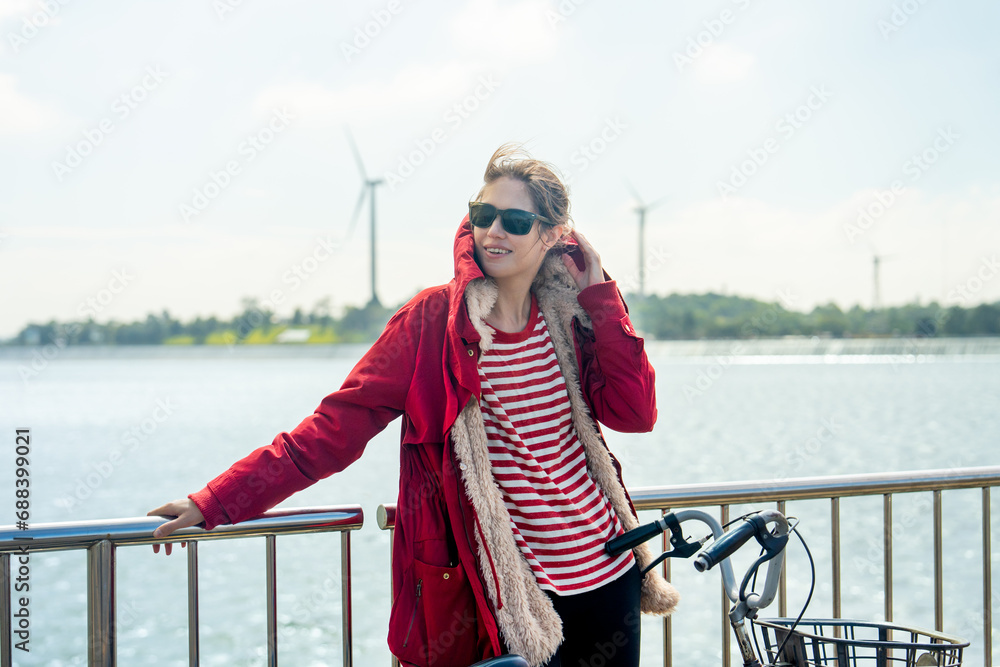 Obraz premium Close up Woman with red coat and sun glasses lean to fence and touch coat hood also hold hand of bicycle stand in front of wind turbines or windmill near water reservoir.