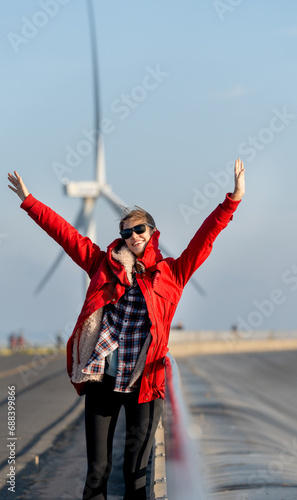 Vertical image of woman with red coat stand with rest her hands up to action of happiness in front of wind turbine or windmill in the back.