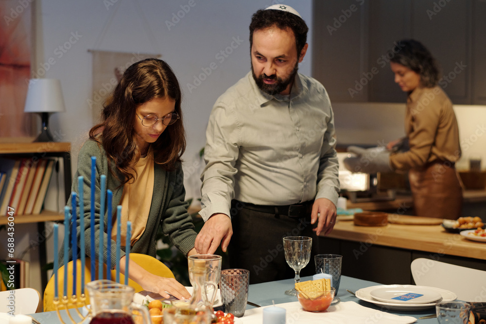 Mature bearded man in kippah standing by his daughter helping him serve table with homemade food and drinks for Hanukkah dinner Mature bearded man in kippah standing by his daughter helping him serve table with homemade food and drinks for Hanukkah dinner