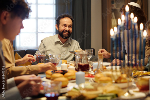 Cheerful mature man looking at one of guests during Hanukkah dinner with his family while enjoying homemade food and drinks photo