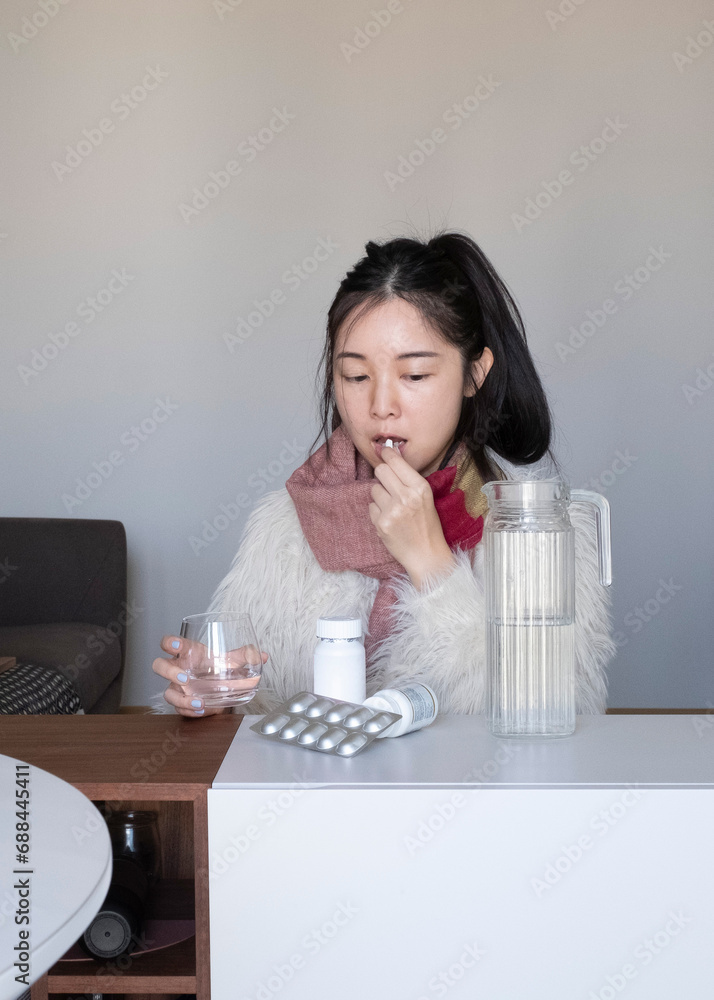 Asian girl taking medicines, pills, supplements at home, in winter clothing and scarf, flu, cold Asian girl taking medicines, pills, supplements at home, in winter clothing and scarf, flu, cold