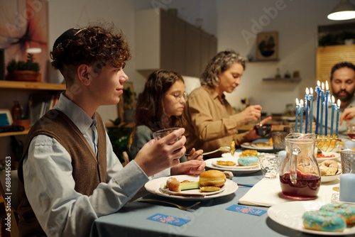 Side view of Jewish boy with curly hair holding glass of homemade drink and having donut and other Hanukkah food during festive dinner