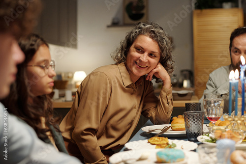 Happy mature Jewish woman looking at one of her children during chat while sitting by table served with homemade food and drinks