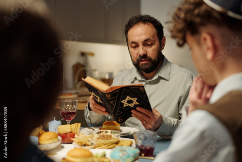 Focus on bearded rabbi and head of Jewish family reading text from Torah while sitting by served table in front of his two children photo