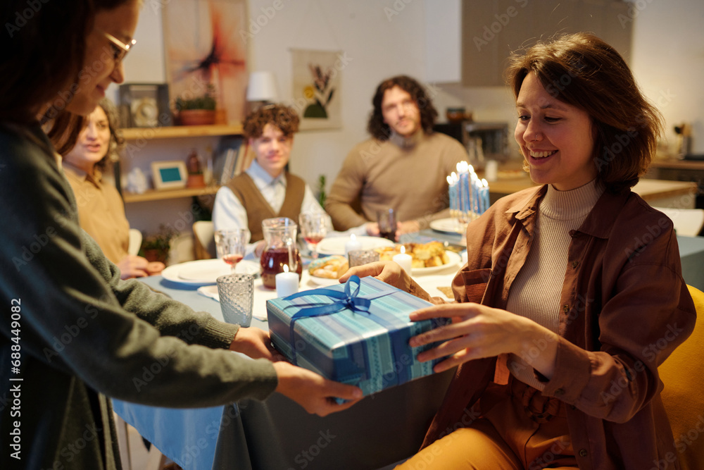 Young smiling woman taking blue giftbox with Hanukkah present from hands of her daughter against other members of family having dinner Young smiling woman taking blue giftbox with Hanukkah present from hands of her daughter against other members of family having dinner