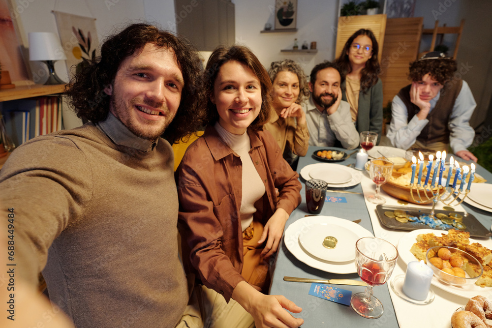 Obraz premium Happy young couple taking selfie with members of their family while sitting by served festive table and enjoying Hanukkah dinner