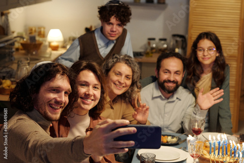 Happy young Jewish man with smartphone taking selfie of large family or communicating in video chat during Hanukkah dinner at home photo