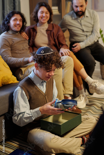 Pre-teen Jewish boy with headphones unpacking giftbox with Hanukkah present from his parents while sitting on the floor by couch photo