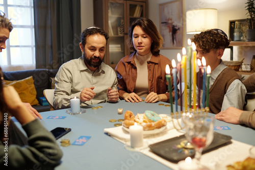 Bearded mature man and his daughter looking at dice while playing leisure game with dreidel, coins and other items after Hanukkah dinner photo
