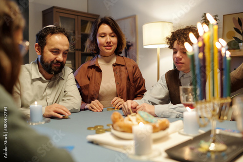 Several members of Jewish family betting during leisure game while sitting by table with burning candles on menorah candlestick photo