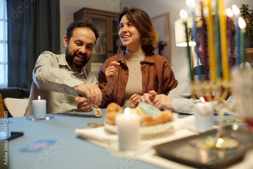 Happy mature man spinning dreidel on table while sitting next to cheerful young woman looking aside during leisure game after dinner photo