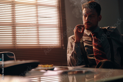 Young serious businessman in formalwear smoking cigarette while sitting by workplace in dark office with window covered with venetian blind