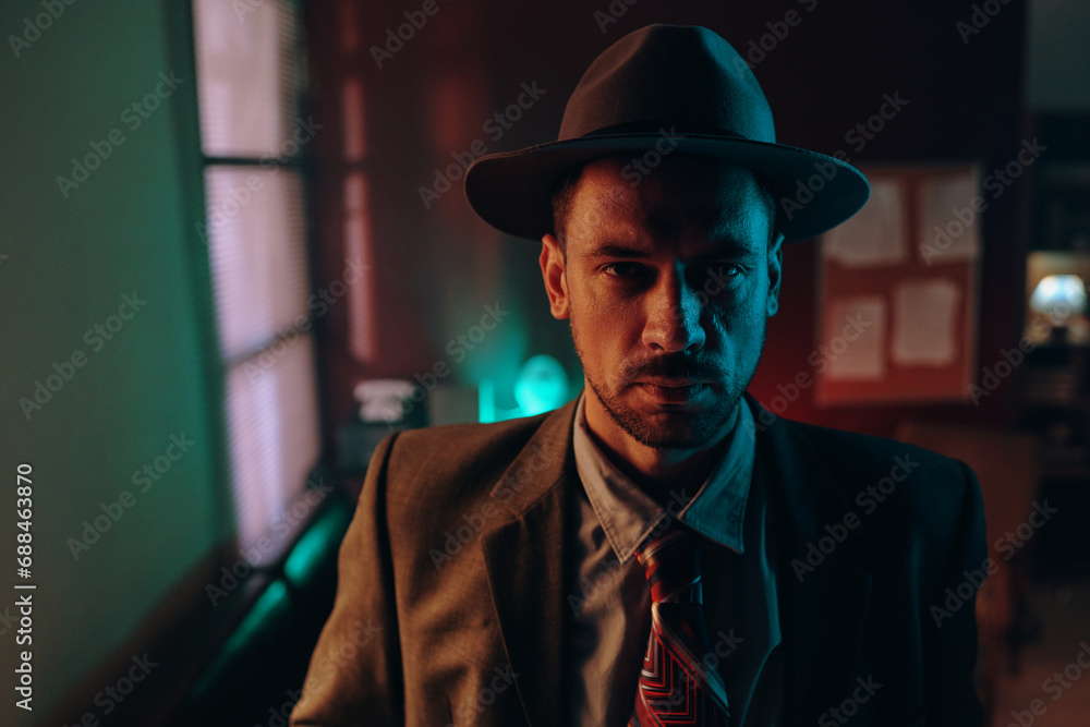 Young serious spy or agent in old-fashioned formalwear and hat looking at you while standing in front of camera in dark office
