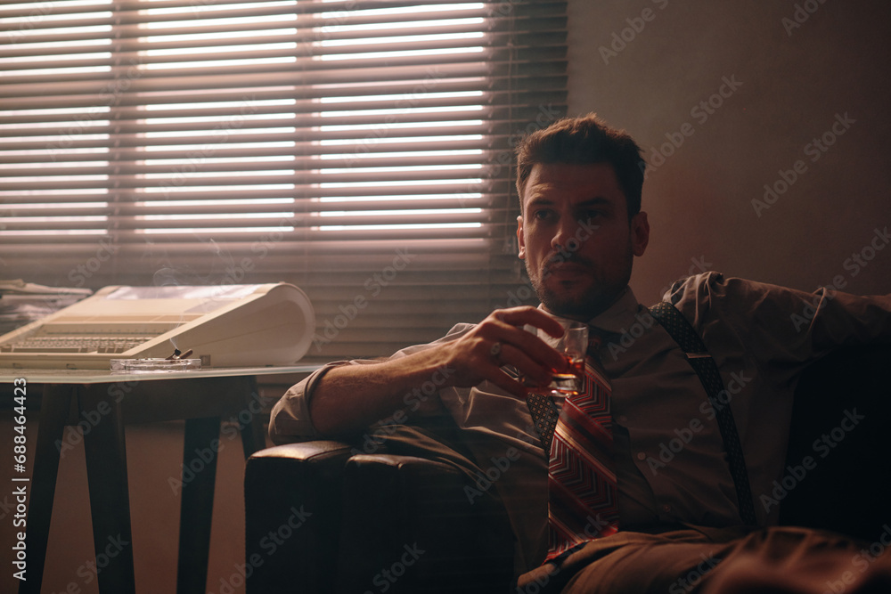 Young serious businessman in retro formalwear holding glass of whiskey while sitting on comfortable couch against window with venetian blinds